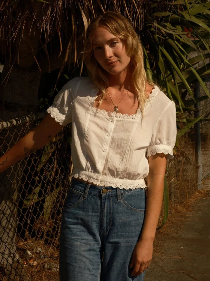 Model wearing Mollusk Surf Shop Lucia Top in white, full front view in warm golden hour light leaning against chain-link fence with tropical foliage, showing square neckline with scalloped eyelet trim, button-front placket, pintuck bodice detail, puff short sleeves with scalloped cuffs, and scalloped eyelet hem, styled with blue high-waist jeans and gold necklace