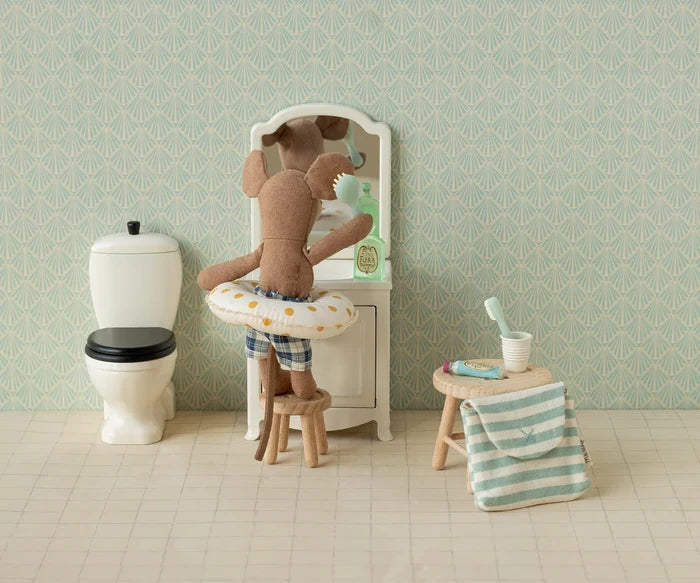 Maileg Toothbrush Set in Blue Striped Bag — lifestyle scene of a Maileg mouse doll in plaid swim trunks brushing teeth at a miniature vanity mirror, with the blue striped bag and toothbrush accessories displayed on a wooden stool nearby