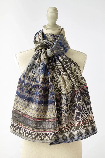 Létol Anoushka Scarf in Jean Trellis, draped and knotted on a linen dress form against a light grey background, showing a reversible Jacquard-woven organic cotton scarf — one side featuring a dense blue and oatmeal floral and trellis pattern with small repeated botanical motifs, the other showing large khaki and oatmeal Mughal medallion and palm tree scrollwork on a taupe ground — with the woven Létol label at the lower center and a pink accent selvedge visible at the hem