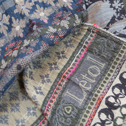 Close-up detail of the Létol Anoushka Scarf in Jean Trellis, showing both sides of the reversible Jacquard weave in layered folds — the denim blue side with oatmeal floral and leaf motifs and coral pink accent threads alongside the khaki and grey side with Mughal scrollwork, botanical bands, and the woven Létol signature, with a bright pink overlock selvedge edge visible between the layers, highlighting the dimensional woven texture and fine detail of the construction