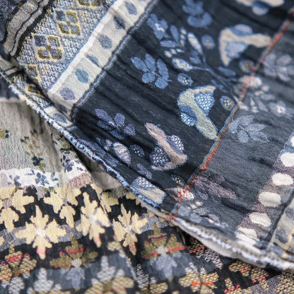 Close-up detail of the Létol Anoushka Scarf in Noir, showing the richly textured Jacquard weave where both sides of the scarf are visible in layered folds — the black ground side with gold and oatmeal oak leaf and floral motifs alongside the oatmeal ground side with chambray blue botanical and paisley patterns, with a red accent selvedge edge visible between the layers, highlighting the dimensional woven construction and tonal contrast between the two sides