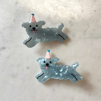 Two blue dog-shaped hair clips with party hats on a marble surface