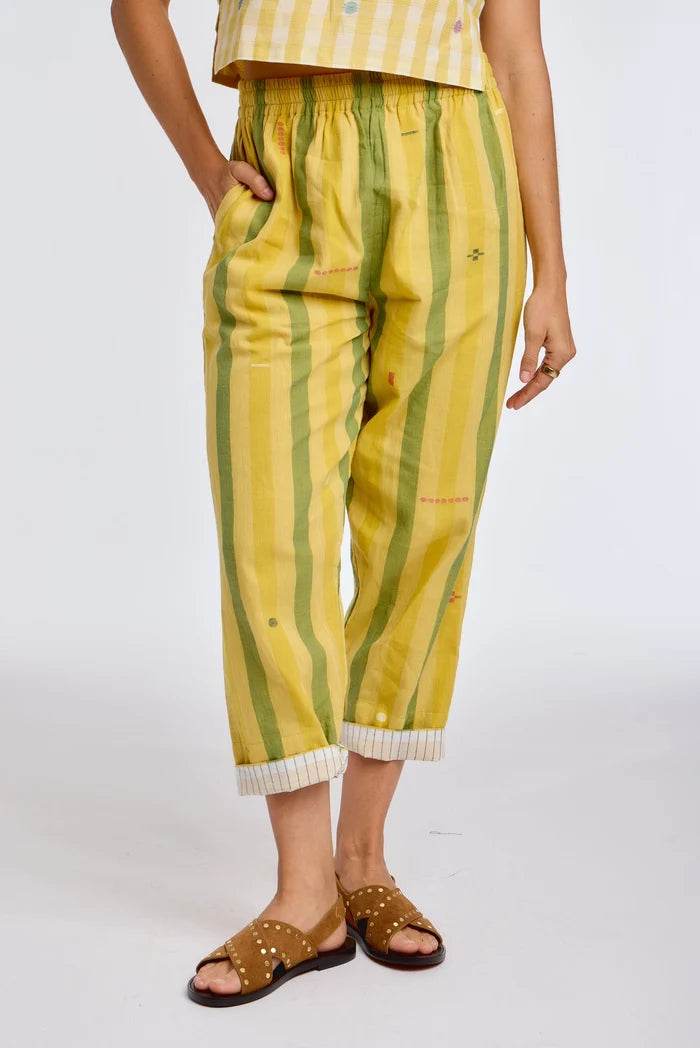 Close-up front view of DVAA Celeste Pant in Goldenrod against white studio backdrop showing goldenrod yellow and sage green wide vertical stripe handwoven cotton with scattered small ikat motifs, elasticated drawstring waistband, hand in side pocket, cropped tapered leg with contrast white pinstripe cuff turn-up, styled with tan gold-studded slide sandals