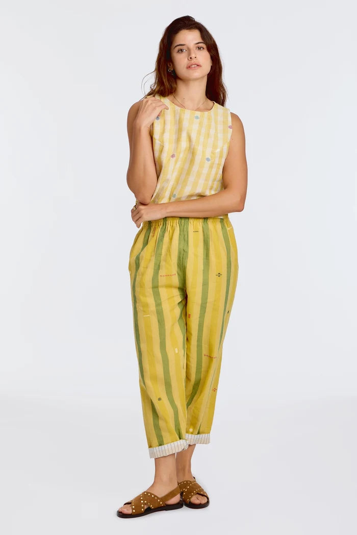 Model wearing DVAA Celeste Pant in Goldenrod, full front view against white studio backdrop showing goldenrod yellow and sage green wide vertical stripe handwoven cotton with scattered ikat motifs, elasticated drawstring waist, cropped tapered leg with contrast white pinstripe cuff turn-up, arms crossed, styled with matching yellow handwoven sleeveless crop top and tan gold-studded slide sandals