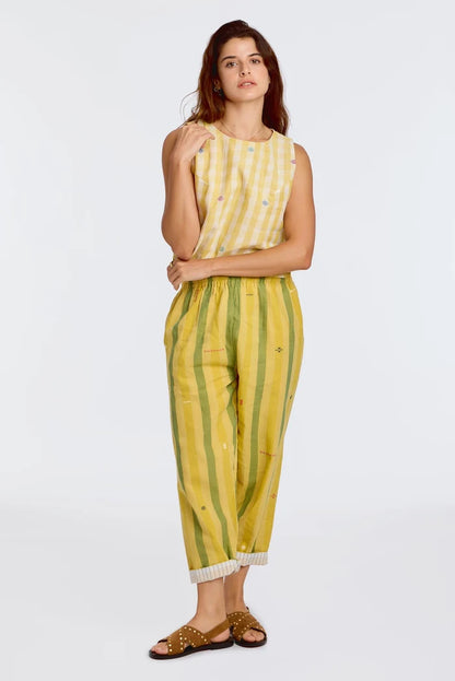 Model wearing DVAA Celeste Pant in Goldenrod, full front view against white studio backdrop showing goldenrod yellow and sage green wide vertical stripe handwoven cotton with scattered ikat motifs, elasticated drawstring waist, cropped tapered leg with contrast white pinstripe cuff turn-up, arms crossed, styled with matching yellow handwoven sleeveless crop top and tan gold-studded slide sandals