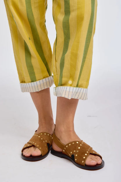 Close-up of DVAA Celeste Pant in Goldenrod hem and footwear detail against white studio backdrop showing goldenrod yellow and sage green wide vertical stripe handwoven cotton, cropped tapered leg with contrast white pinstripe cuff turn-up, styled with tan leather gold-studded crossband slide sandals