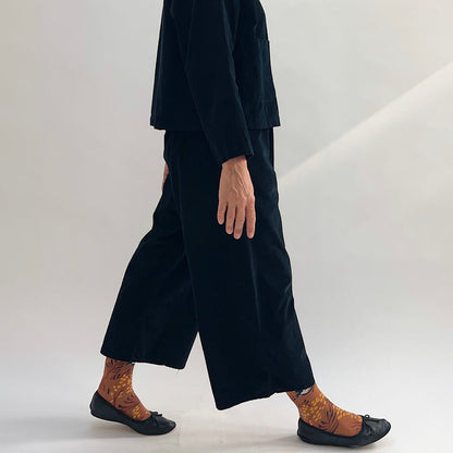 Model walking in the Bryn Walker Corduroy Flood Pants in Black, side profile showing wide-leg silhouette, flood-length cropped hem, and black pinwheel corduroy, paired with matching jacket, printed socks, and black ballet flats