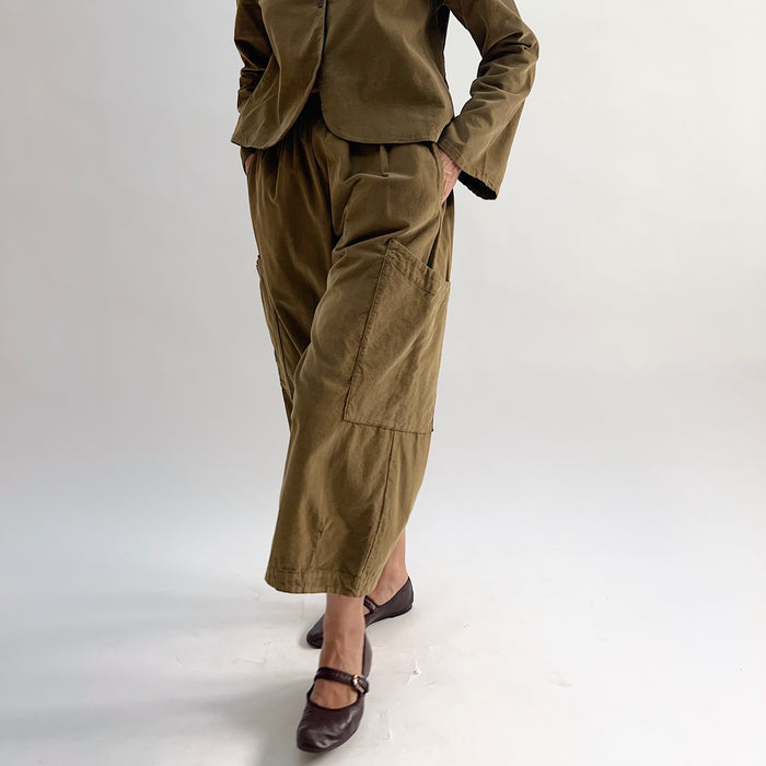 Model with hand on hip wearing the Bryn Walker Corduroy Pasha Pants in Partridge, front view showing side leg patch pockets, elastic waistband, and olive pinwheel corduroy, paired with matching Audrey Jacket and dark Mary Jane flats