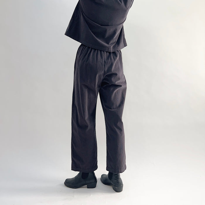 Back view of model wearing the Bryn Walker Corduroy Pleated Pants in Fig, showing elastic back waistband, tapered straight leg silhouette, and deep fig pinwheel corduroy drape, styled with matching Standing Collar Crop Jacket and black Chelsea boots