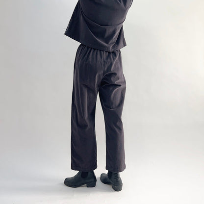 Back view of model wearing the Bryn Walker Corduroy Pleated Pants in Fig, showing elastic back waistband, tapered straight leg silhouette, and deep fig pinwheel corduroy drape, styled with matching Standing Collar Crop Jacket and black Chelsea boots