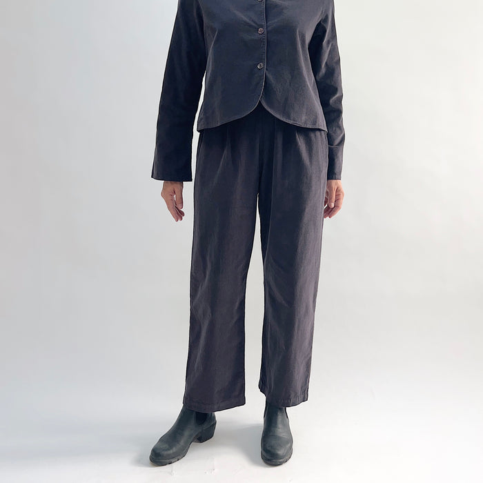 Model wearing the Bryn Walker Corduroy Pleated Pants in Fig, full front view showing flat front with pleats, elastic waistband, tapered straight leg, and deep fig pinwheel corduroy, styled with matching Standing Collar Crop Jacket and black Chelsea boots