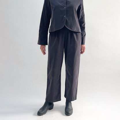 Model wearing the Bryn Walker Corduroy Pleated Pants in Fig, full front view showing flat front with pleats, elastic waistband, tapered straight leg, and deep fig pinwheel corduroy, styled with matching Standing Collar Crop Jacket and black Chelsea boots