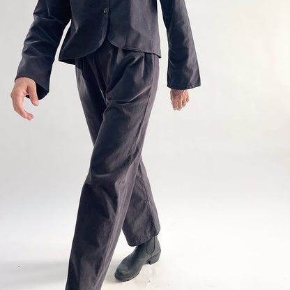 Model in motion wearing the Bryn Walker Corduroy Pleated Pants in Fig, close-up showing pleated flat front, tapered straight leg movement, slit hem detail, and deep fig pinwheel corduroy, styled with matching Standing Collar Crop Jacket and black Chelsea boots