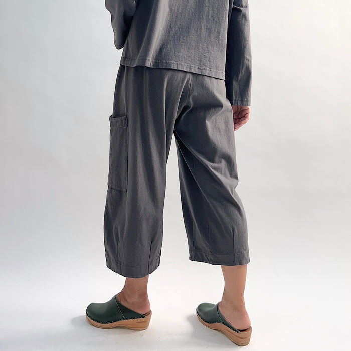 Back view of model wearing the Bryn Walker Cotton Pasha Pant in Moonlight, showing elastic gathered waistband, wide-leg cropped silhouette, thigh patch pocket, folded cuff hem, and soft blue-grey cotton, styled with matching top and green wedge clogs