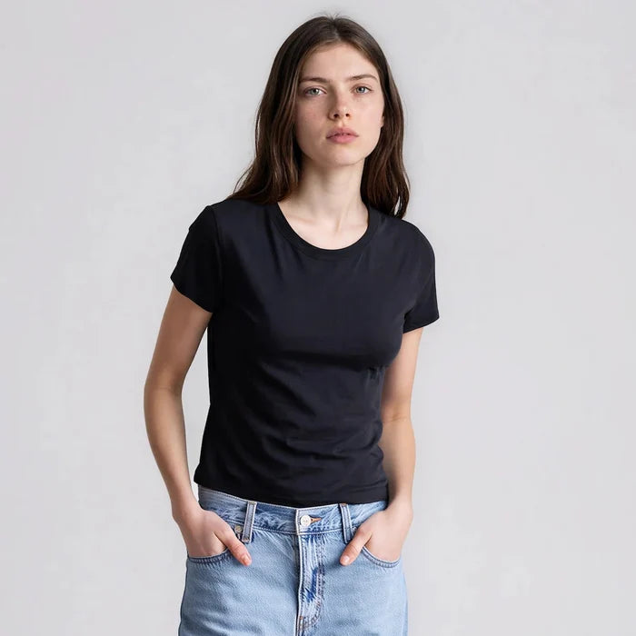 Model wearing It Is Well Cropped Tee in Black, front view against light grey background showing solid black lightweight slub jersey, clean crew neck, short fitted sleeves with slight cuff, and slightly cropped length in slim petite fit, styled with light wash high-rise jeans, hands in front pockets