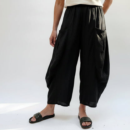 A person wearing loose, wide-leg pleated pants in a light beige and gray crinkled fabric, paired with a beige top and black slide sandals, photographed against a plain white studio background.