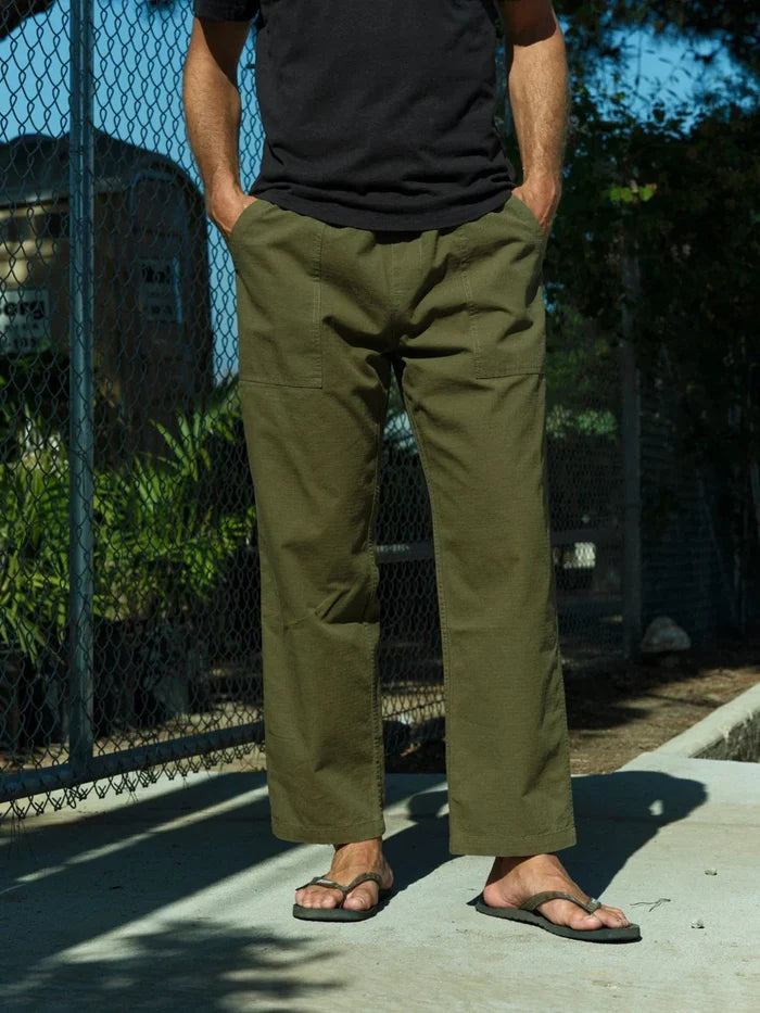 Model wearing Mollusk Surf Shop Drawstring Salvador Pants in Mash Green Ripstop, front view showing olive green double-washed cotton ripstop, roomy straight-leg silhouette, elastic drawstring waistband, front slash pockets, and utility carpenter panel seam detail on thigh, styled with black t-shirt and olive flip flops outdoors near chain-link fence