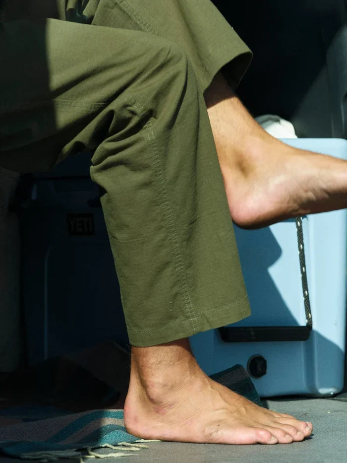 Close-up lifestyle shot of Mollusk Surf Shop Drawstring Salvador Pants in Mash Green Ripstop showing relaxed ankle hem drape and double-washed olive green cotton ripstop fabric texture, model barefoot stepping on a cooler in an outdoor van setting