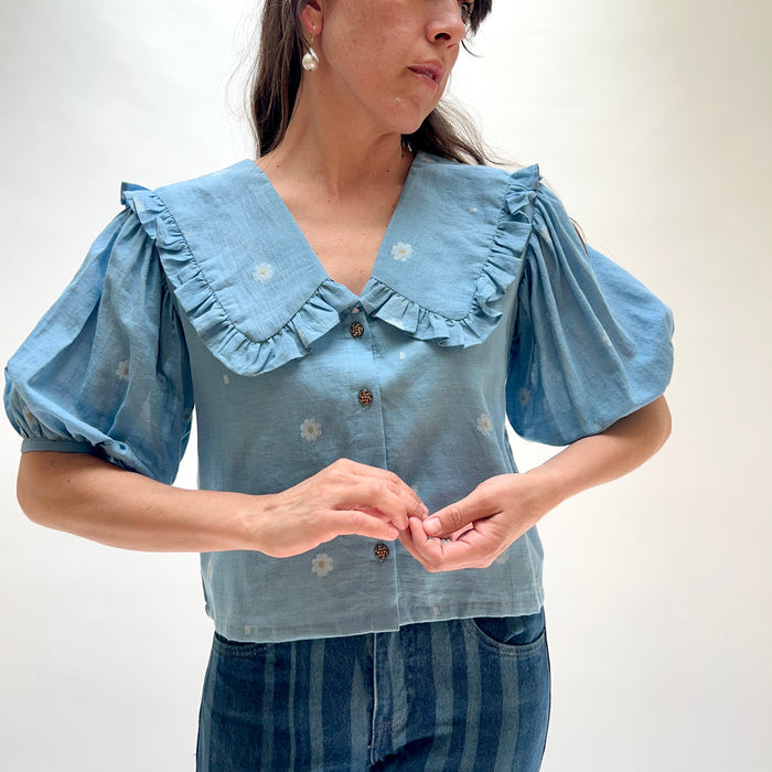 Model wearing DVAA Fleurie Blouse in Aurelia Blue, front cropped view against white studio backdrop showing dusty blue 100% cotton woven daisy jacquard, oversized ruffled peter pan collar with V-neckline, voluminous puff short sleeves, and dark tortoiseshell button-front placket, styled with dark blue striped wide-leg jeans and pearl drop earrings
