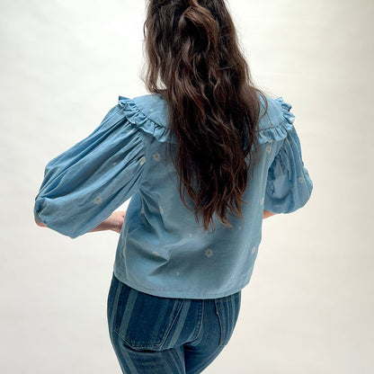 Model wearing DVAA Fleurie Blouse in Aurelia Blue, back view against white studio backdrop showing dusty blue 100% cotton woven daisy jacquard, oversized ruffled peter pan collar, voluminous puff short sleeves with elasticated cuffs, and cropped boxy silhouette, styled with dark blue striped wide-leg jeans