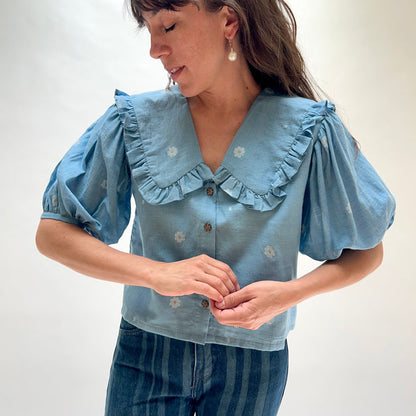 Model wearing DVAA Fleurie Blouse in Aurelia Blue, full front view against white studio backdrop showing dusty blue 100% cotton woven daisy jacquard pattern, oversized ruffled peter pan collar with V-neckline, voluminous puff short sleeves with elasticated cuffs, and dark tortoiseshell button-front placket, styled with dark blue striped wide-leg jeans and pearl drop earrings