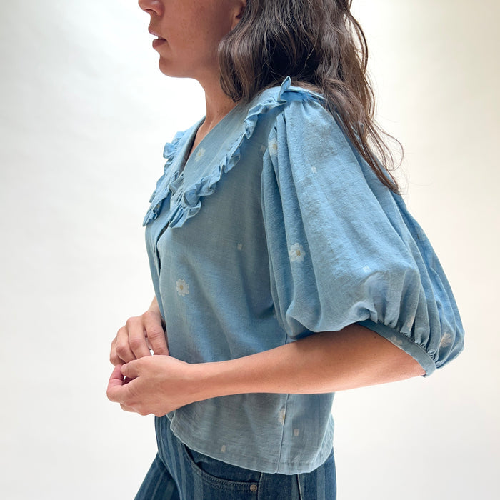 Model wearing DVAA Fleurie Blouse in Aurelia Blue, side profile view against white studio backdrop showing dusty blue 100% cotton woven daisy jacquard, oversized ruffled peter pan collar, voluminous puff short sleeve with elasticated cuff, button-front placket, and cropped boxy fit, styled with dark blue jeans