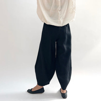 Back view of model wearing the Bryn Walker Heavy Linen Oliver Pant in Black, highlighting barrel-leg volume, elastic waistband, and tapered ankle in black heavy linen, styled with cream linen top and black ballet flats