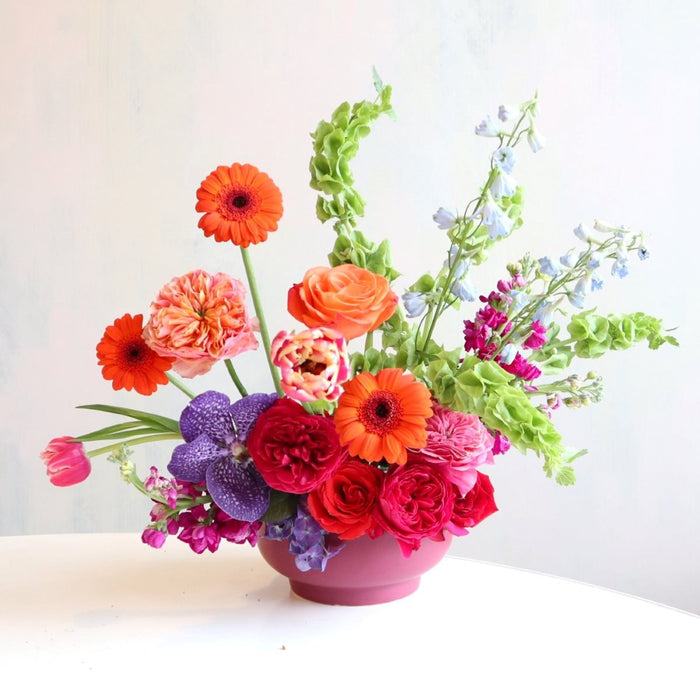Bright and colorful floral arranegement, with orange gerbera daisies, bells of ireland, fushia garden roses, blue delphinium, purple vanda orchids, burgundy stock in a purple low compote.