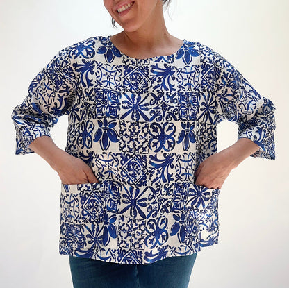 Front view of the Christopher Calvin Linen Blue Tiles Print Top with hands in the two front patch pockets, showing the relaxed silhouette, boat neckline, and blue-and-white tile print, on a white background