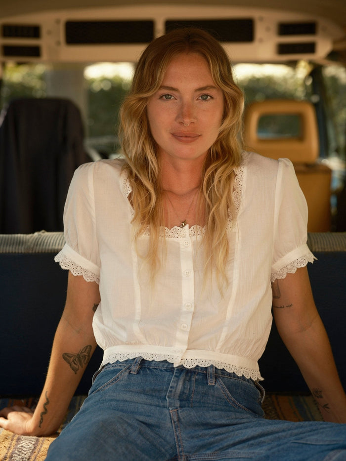 Model wearing Mollusk Surf Shop Lucia Top in white, seated inside a vintage van, front view showing square neckline with scalloped eyelet trim, button-front placket with pintuck bodice detail, puff short sleeves with scalloped eyelet cuffs, and scalloped eyelet hem, styled with blue high-waist jeans and gold necklace