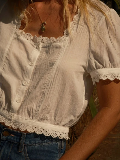 Close-up of Mollusk Surf Shop Lucia Top in white showing square neckline with scalloped eyelet trim, button-front placket with pintuck bodice detail, puff short sleeves with scalloped eyelet cuffs, and scalloped eyelet hem on 100% cotton airy fabric, styled with blue jeans and gold pendant necklace in warm outdoor light