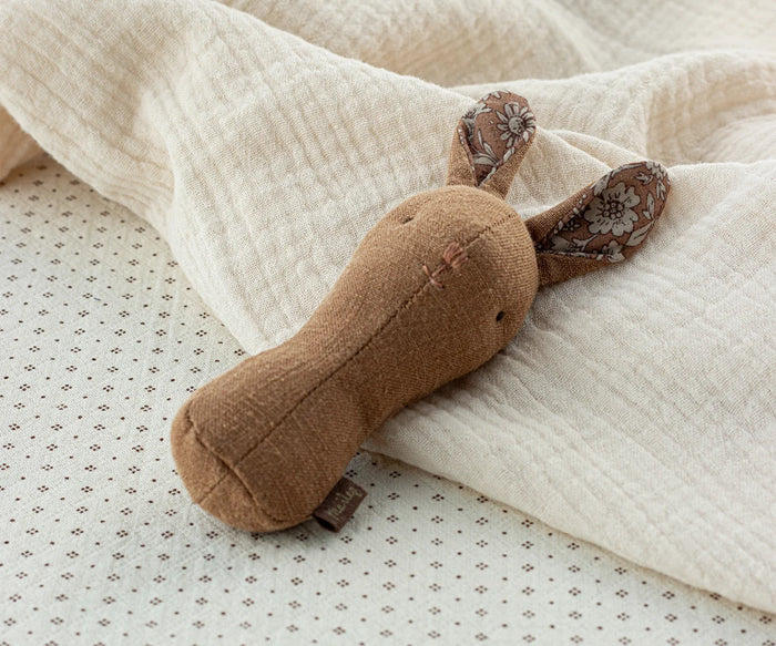 Maileg Lullaby Friends Bunny Rattle in Chocolate resting against a soft cream muslin blanket — a warm brown linen bunny rattle with floral-lined ears