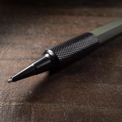 A close-up of a mechanical pencil with a textured black grip and a dark metallic tip, resting on a dark wooden surface.