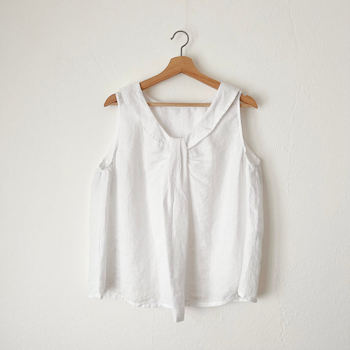 A white linen sleeveless top with a round neckline, hanging on a wooden hanger against a white wall.
