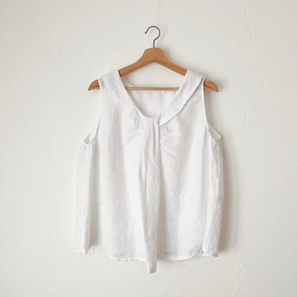 A white linen sleeveless top with a round neckline, hanging on a wooden hanger against a white wall.