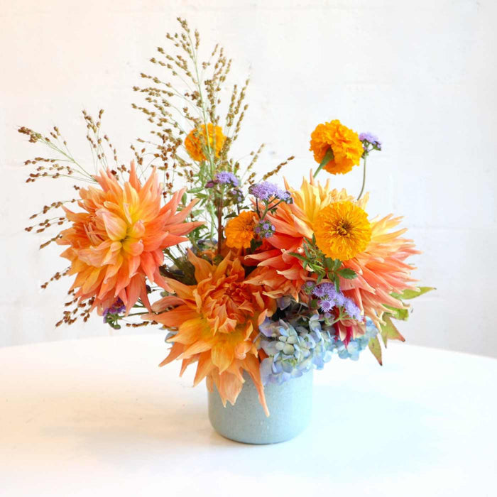 Arrangement in blue vase with orange dahlias, orange marigolds, purple ageratum, fall grass on a white background.