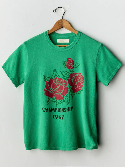 A green short-sleeve t-shirt hanging on a wooden hanger against a light gray wall. The shirt features a graphic print of three red roses with green leaves and the text 'CHAMPIONSHIP 1967' in black below the roses.