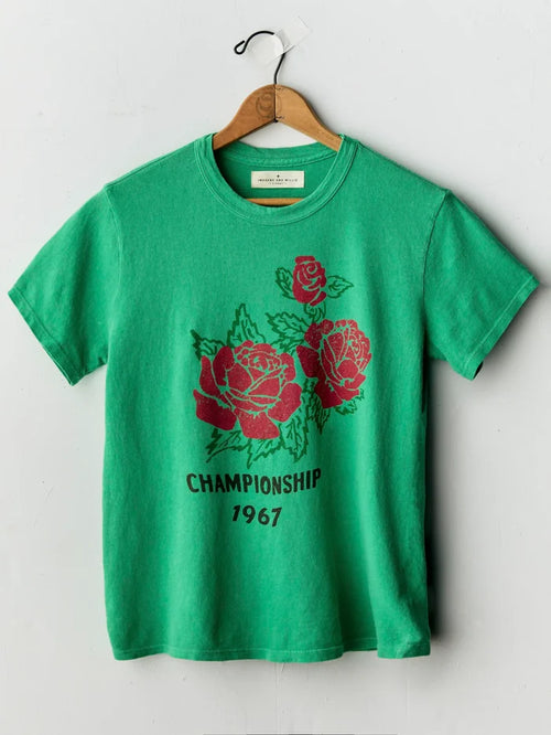 A green short-sleeve t-shirt hanging on a wooden hanger against a light gray wall. The shirt features a graphic print of three red roses with green leaves and the text 'CHAMPIONSHIP 1967' in black below the roses.