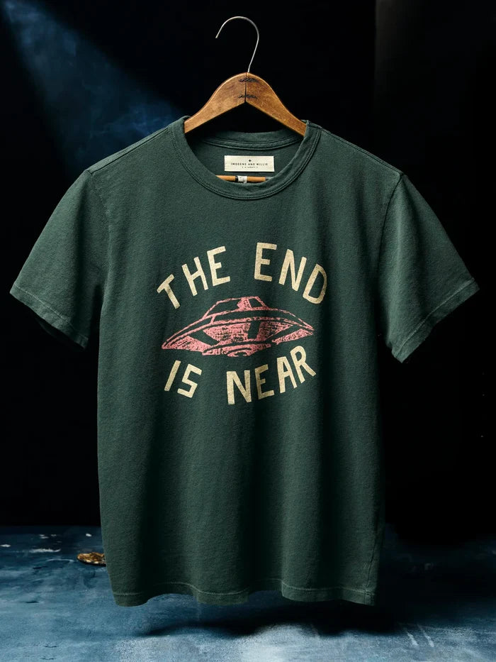 Dark spruce green short-sleeve tee on a wooden hanger, screen-printed with "The End Is Near" text and a vintage UFO graphic in cream and pink.