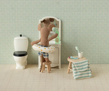 Maileg Toothbrush Set in Blue Striped Bag — lifestyle scene of a Maileg mouse doll in plaid swim trunks brushing teeth at a miniature vanity mirror, with the blue striped bag and toothbrush accessories displayed on a wooden stool nearby