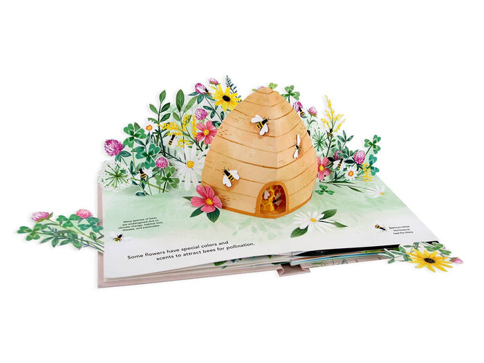 Flora: A Botanical Pop-Up Book | Open spread showing a beehive and floral garden. Text reads "Many species of bees are endangered due to climate change, habitat loss, disease, and pesticides. Some flowers have special colors and scents to attract bees for pollination. Bees to nectar into honey to feed the colony.".