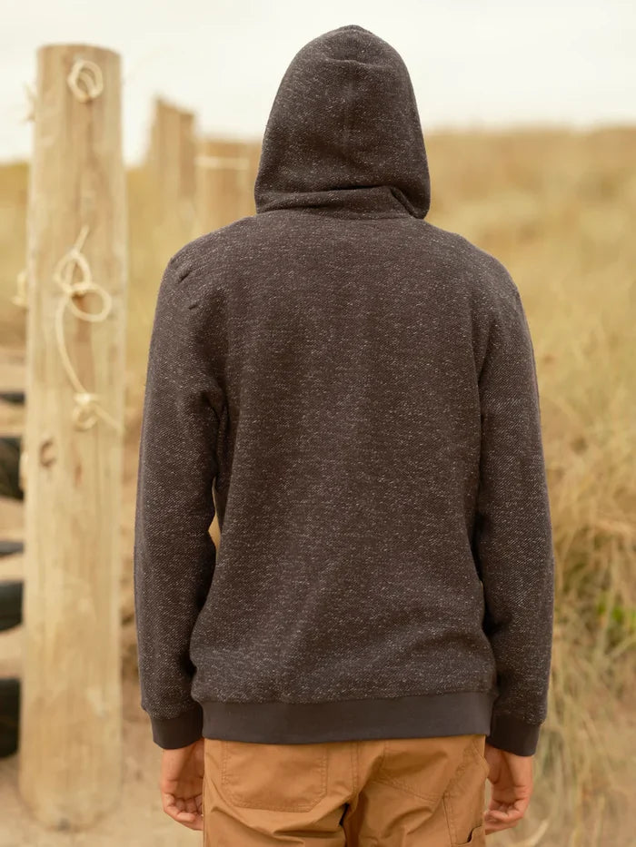Back view of male model wearing a faded navy garment-dyed zip-up hoodie with hood up, showing the textured terry knit fabric, relaxed fit, and ribbed hem and cuffs, styled with tan pants against a coastal dune background.